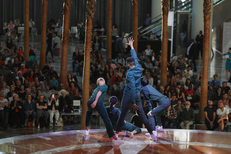 Four performers in everygreen-colored jumpsuits pose and look down at the audience.